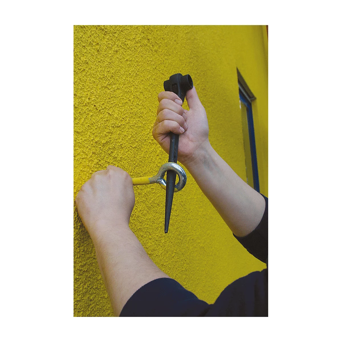 Hände halten Werkzeug zur Wandmontage. Gelbe Wand im Hintergrund, Fenster. Werkzeug: Schwarz, Gelb und Silber.